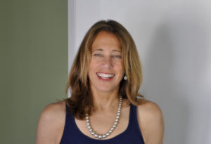 Susan Kare, a handsome fifty year old woman with dark blonde hair, a tan complexion, and green eyes, smiles broadly. She is wearing a purple tank top and a pearl necklace