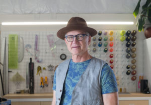 Tinker is in a design studio. A white man in his sixties or seventies, he wears a hip brown fedora, cool glasses, a nice patterned shirt, and a blue vest.
