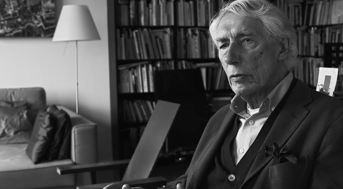 A black and white portrait of Wim Crouwel seated in a study. He looks dignified. He gazes into the near distance. He is wearing a suit with no tie. He has sweeping white hair.