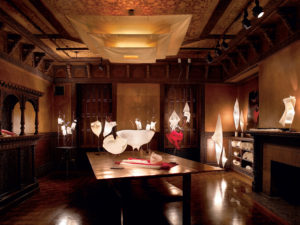 In the ornate, wood-paneled Teak Room of Cooper Hewitt, a dozen illuminated white sculptures cast a warm glow. The sculptures look like sheets of fabric suspended and spinning in mid-air. There are also two of these forms in red. On a the corner are process models that make clear these ethereal forms are actually cast in plaster. Three gold sheets are suspended in layers near the top of the ceiling with a gold light projected through them. This creates an additional source of illumination.