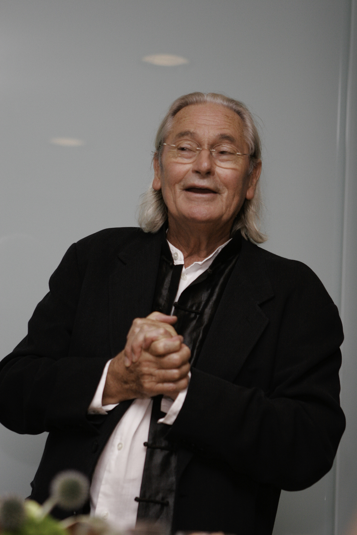 Ingo smiles widely and clasps his hands in delight. He is an older white gentleman with shoulder-length silver hair. He is wearing a black jacket and a white button-up shirt.