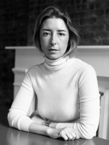 black and white photograph of Alexandra Cunningham Cameron, curator of contemporary design at Cooper Hewitt. She is seated at a table with her arms folded in front of her. She wears a white turtleneck.