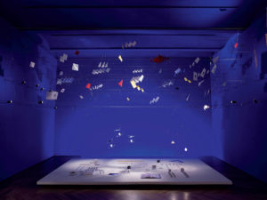 A room with a wooden floor has a wall and ceiling that are painted indigo. About two dozen thin black wires criss-cross the room, like electricity cables. Suspended from these wires are whimsical assortments of square cut outs in yellow, red, and translucent materials. Dangling further down are small lightbulbs. On the floor is a white platform on which are a book and lighting components neatly organized.