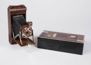 Image features leather covered folding camera with front panel hinged to open and reveal a bellows with lens housing enameled in red, brown and tan geometric pattern. Rectangular black-lacquerd cedar box with same geometric design as on camera holds the folded camera. Please scroll down to read the blog post about this object.