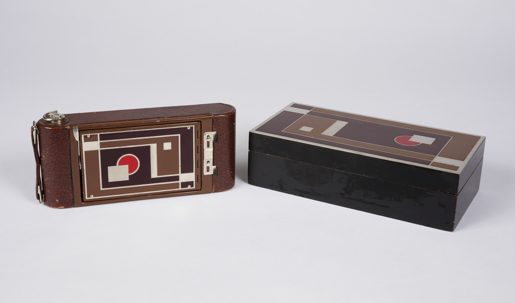 Image features leather covered folding camera with front panel enameled in red, brown and tan geometric pattern. Rectangular black-lacquerd cedar box with same geometric design as on camera holds the folded camera. Please scroll down to read the blog post about this object.