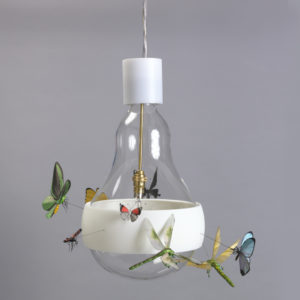 Hanging lamp in the form of a large clear glass incandescent light bulb, the globe surrounded by a transluscent white plastic band from which project eight colorful, realistic figures of winged insects seemingly in flight around the bulb; halogen light source suspended from brass fitting in center of bulb-shaped globe.