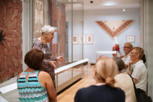 A group of people in a brightly lit gallery with red textiles hanging on all of the walls. A guide leads a discussion