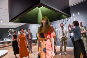 Interpretation as Storytelling. A woman in a colorful outfit stands under a large grey box installed from the ceiling and looks up. Behind her, a group of people watch within the gallery space. Below is program information including date and ticketing.