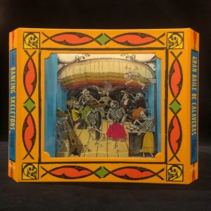 Image features the cover of the book, Gran Baile de Calaveras, un Libro Túnel, a yellow, orange, and green rectangular frame surrounding an image of a gathering of skeletons in brightly hued clothing dancing, socializing, and celebrating. Please scroll down to read the blog post about this object.