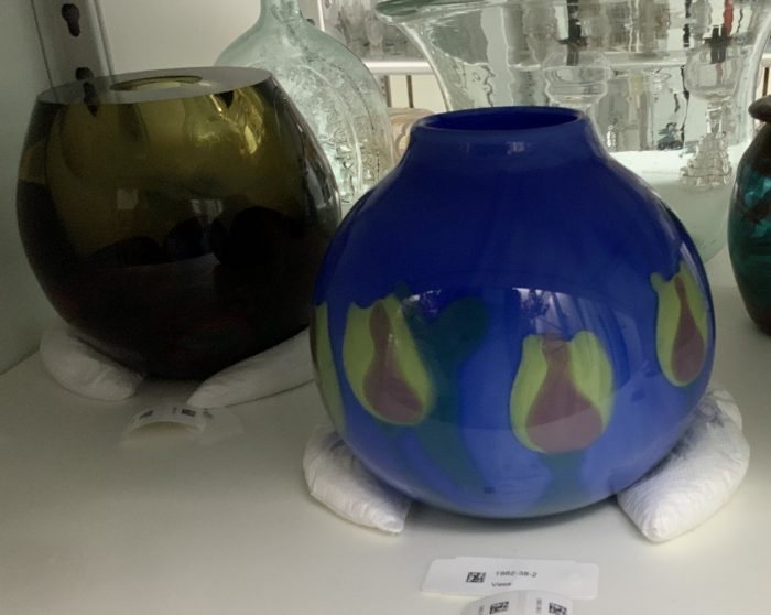 Two glass bowls, one dark green and one blue with green, flower-shaped imagery, are on a shelf. White, padding materials are gathered around their bases to hold them in place.