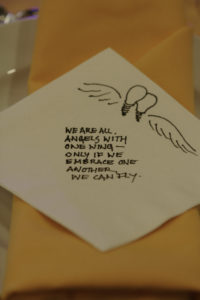 A napkin on which there is a sketch of two lightbulbs nestled together. Each lightbulb has one wing, and they seem to be helping one another soar. Text: We are all angels with one wing - only if we embrace one another can we fly."