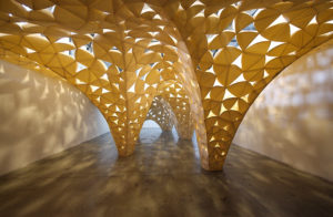 Light streams through a golden architectural structure. The structure is similar to a canopy roof that extends in three pillars to touch the ground. The structure is deliberately punctured with triangle-shaped holes.