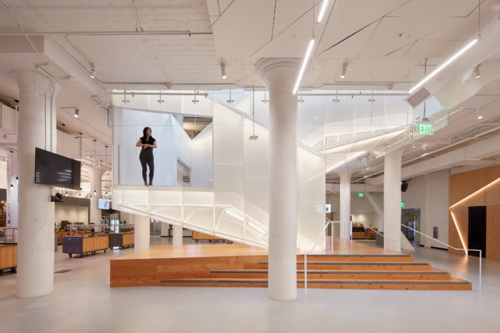 An airy open floor office space toes the line between the modern and the futuristic. A woman in dark clothing looks at the room through a mezzanine in a stairwell seemingly constructed from beams of light. A graceful wooden staircase leads to the stairwell. In the background and on the ceiling are architectural details, like robust pillars and an exposed HVAC system, that suggest this building was once an industrial warehouse.