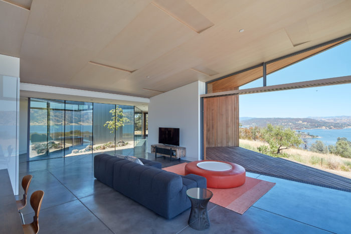 A tranquil modern sea front home with a living room with a huge glass door that opens to unobstructed views of a sloping grassy hill, a tranquil body of water, and distant mountains