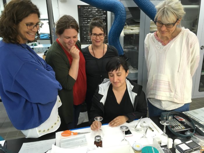 Five people in a lab circle a table and look at an upside down wine glass. Other objects, such as scissors, jars, and pens, are located on the table.