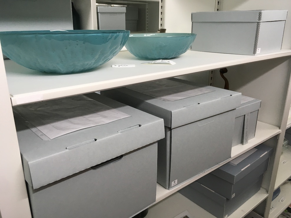 Photograph of three shelves in a storage facility. All shelve archival gray boxes, and the top shelf also holds two teal glass bowls.