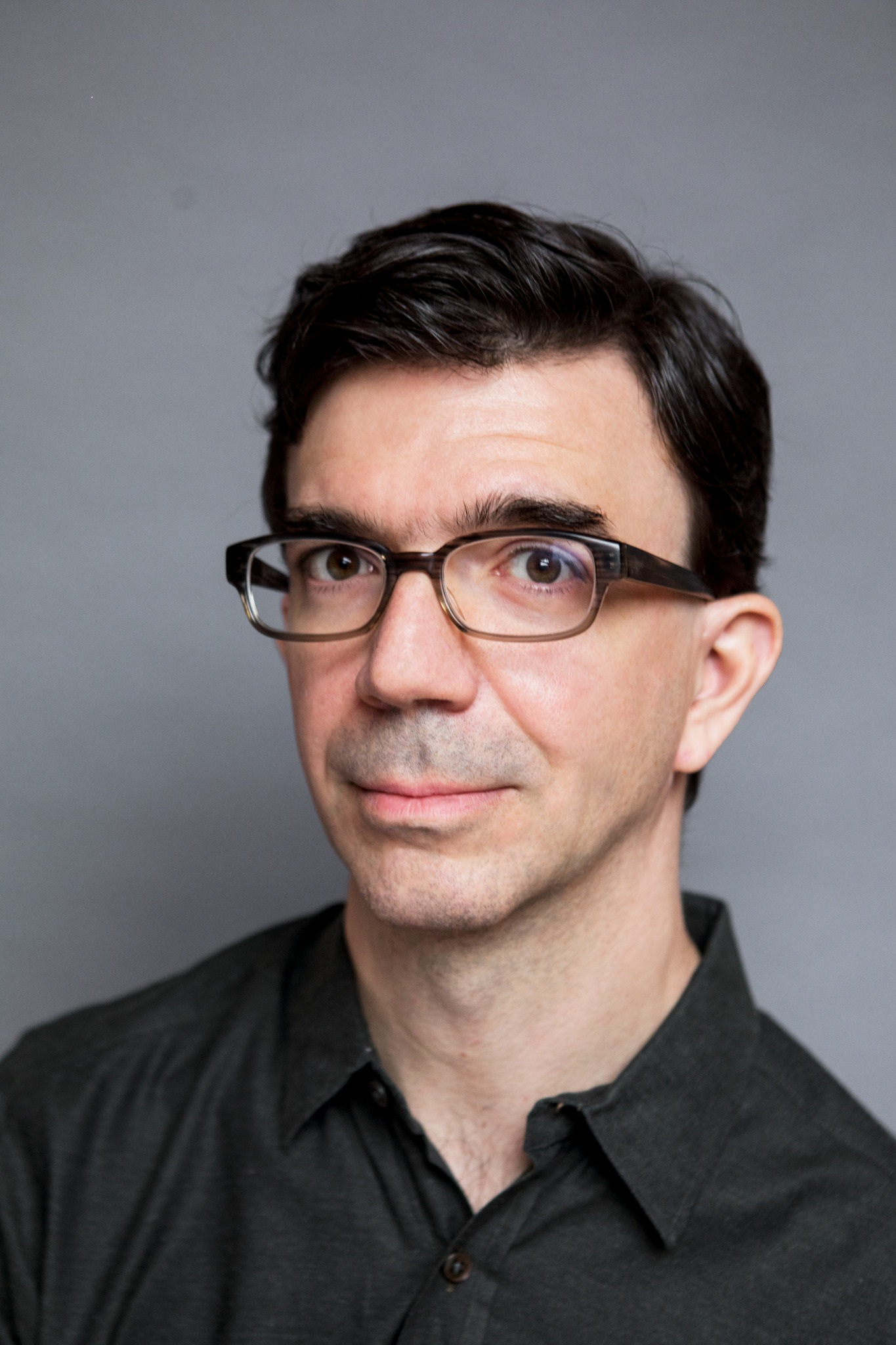 Tobias, a white man around forty with dark hair, dark eyes, wearing black-framed glasses and a black polo shirt. He's slightly smiling with his mouth closed.