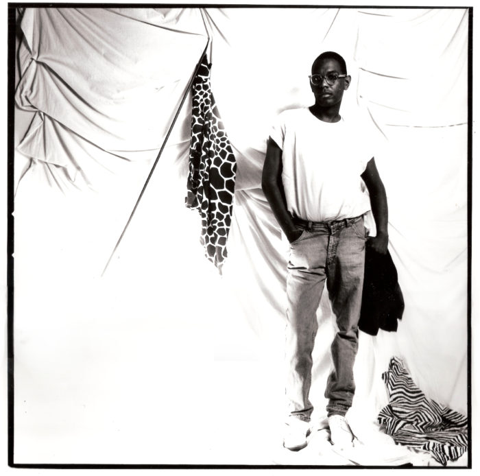 A portrait of Willi Smith, a young African American man wearing a white t-shirt, jeans, and spectacles, in a photo studio. Behind him is draped a white sheet. A small piece of giraffe-print fabric is draped from a leaning flag pole. At his feet is a zebra-print blanket or piece of fabric thrown haphazardly on the ground.