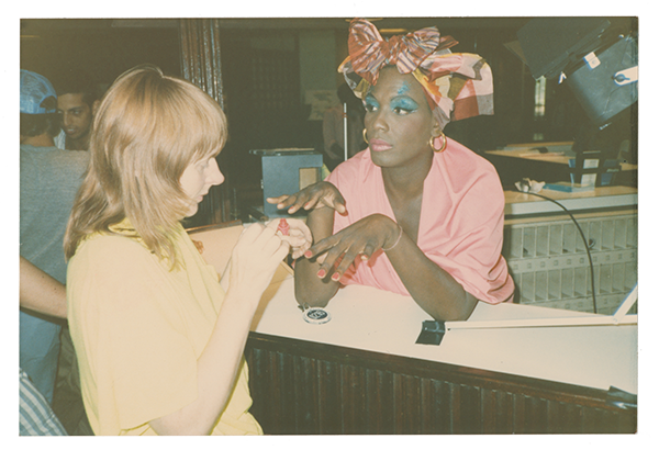 Photograph of a woman in yellow painting Willi Smith's nails, who is wearing a pink blouse, makeup, and a head wrap