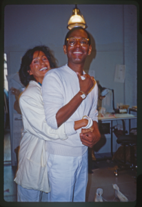 Photograph of a woman hugging Willi Smith from behind, both wearing white and smiling at the camera