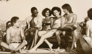 Black and white image of a group of people, including Willi Smith, smiling and seated by a pool.