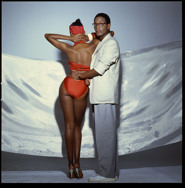 Willi Smith facing the camera standing with a woman in a red bikini and metallic heels, whose back is to the camera
