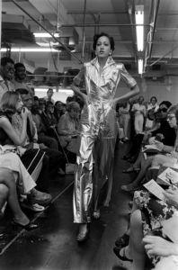 Black and white photograph of a model in a fashion show wearing a reflective jumpsuit