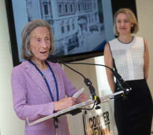 Barbara speaks at a podium while Caroline Baumann looks on with admiration and respect