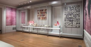 Gallery view showing a selection of textiles and objects on display, with pink variegated wallpaper surrounding the room.