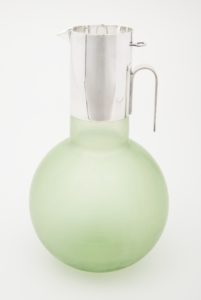 Image features a pitcher composed of a globular, translucent green glass body with a cylindrical neck covered in silver-plated metal with an inverted U-shaped handle, short spout, and an inset circular lid. Please scroll down to read the blog post about this object.