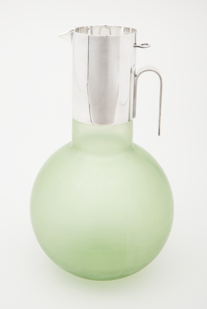 Image features a pitcher composed of a globular, translucent green glass body with a cylindrical neck covered in silver-plated metal with an inverted U-shaped handle, short spout, and an inset circular lid. Please scroll down to read the blog post about this object.