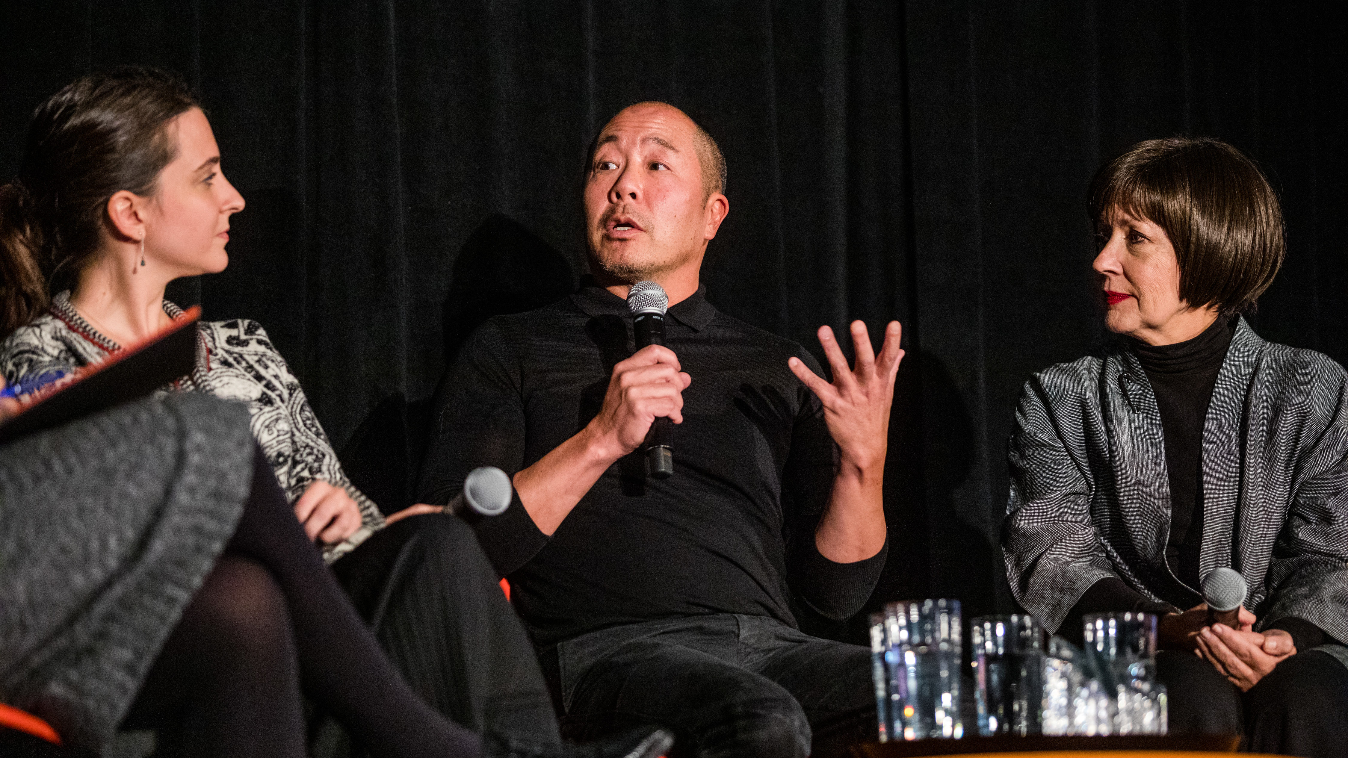 Image of Pinar Guvenc, Derek Lam and Patricia Moore, 2019 NDA winners, sitting together on stage, all in dark grey and black clothing, with a dark background, Derek holds a microphone with one hand and gestures with the other