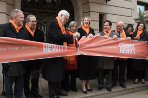 A group of people in winter coats wearing orange scarfs cut a big orange ribbon in front of Carnegie Mansion. Among the people in the photo are Barbara and Caroline Baumann