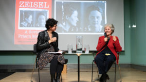 Image of two women sitting in chairs on a stage. Both have microphones. Left: Margaret Gould Stewart, who has short black curly hair, black leather jacket, and a patterned skirt. She wears glasses. She appears to be in her 40s. Right: Jean Richards, who has short curly grey hair, a red cardigan and black pants. She appears to be in her 70s and speaks into the microphone with a smile.