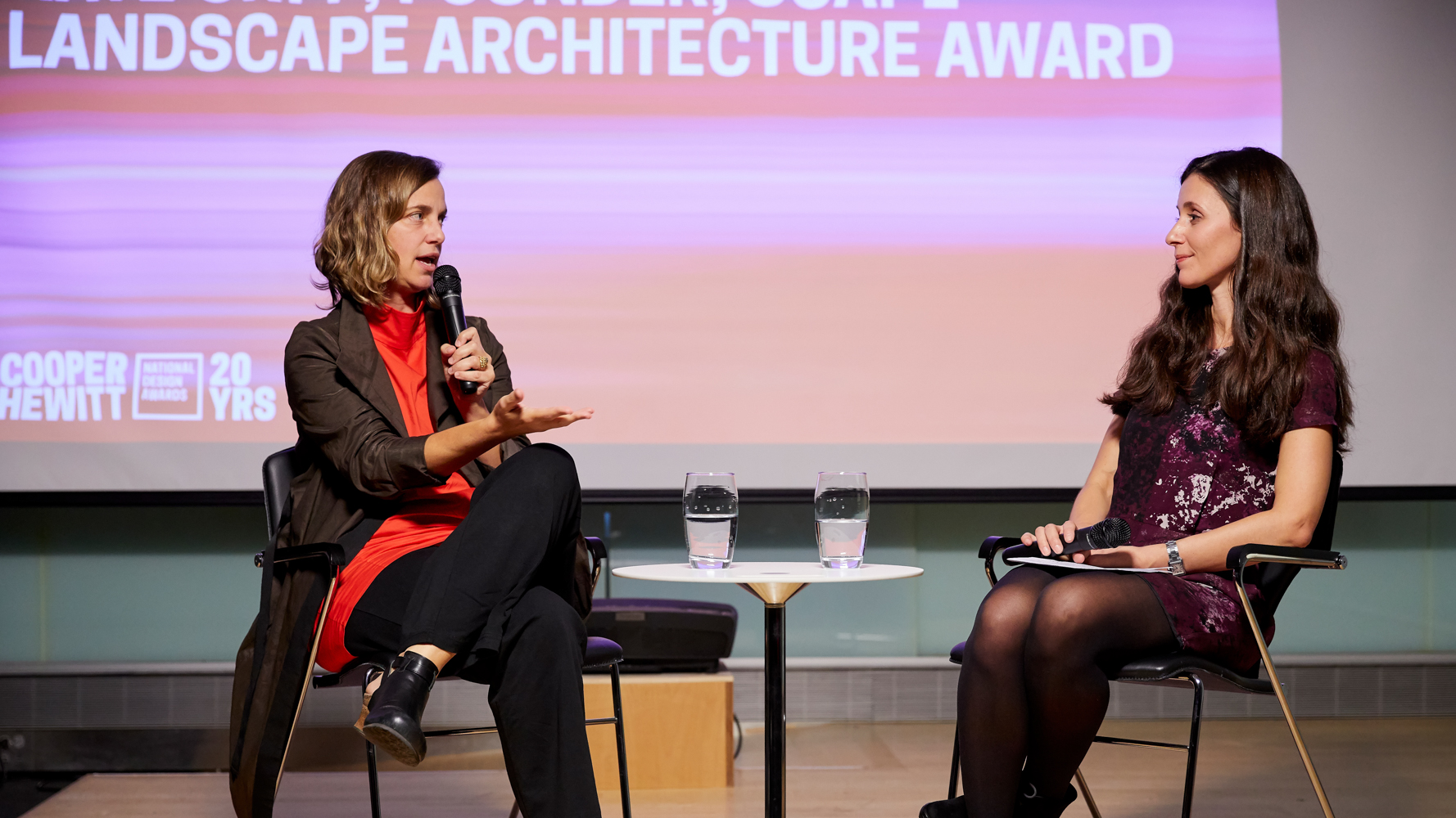 image of two women on stage, on the left is Kate Orff, wearing a red blouse under a brown suit jacket, she holds a microphone in her left hand, speaking into it, and gestures with her right hand. her hair is brown and above the shoulders. ON the left is Vasso Giannopolis who wears a dark purple dress and has long brown hair. She looks and listens.