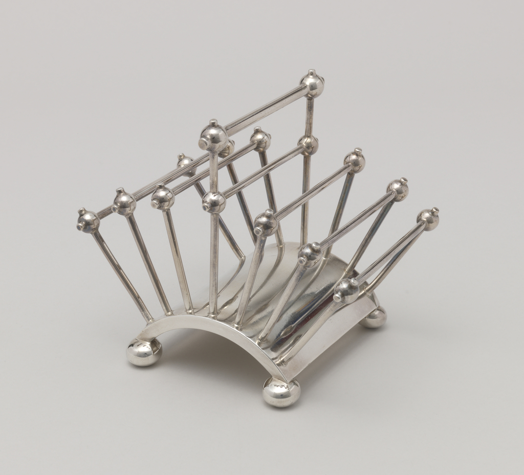 Image features a silver-plated toast rack with seven square dividers composed of vertical posts and horizontal rods connected by ball joints, the center one with an extra raised section to serve as a handle, all on a curved base resting on four bun feet. Please scroll down to read the blog post about this object.