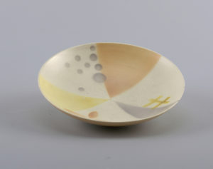 Image features a circular dish, the surface with airbrushed geometric decoration of pale peach, grey and yellow triangles and small circles on a cream-colored ground. Please scroll down to read the blog post about this object.