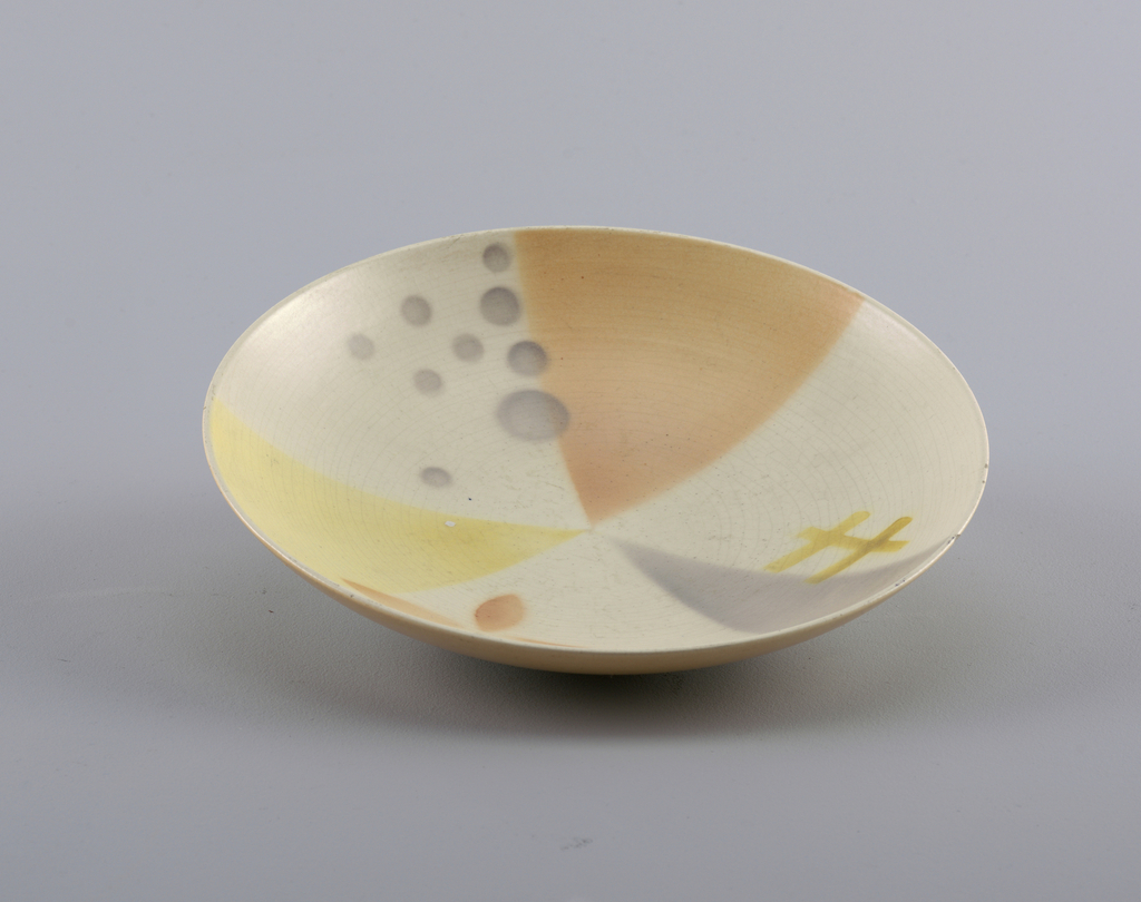 Image features a circular dish, the surface with airbrushed geometric decoration of pale peach, grey and yellow triangles and small circles on a cream-colored ground. Please scroll down to read the blog post about this object.