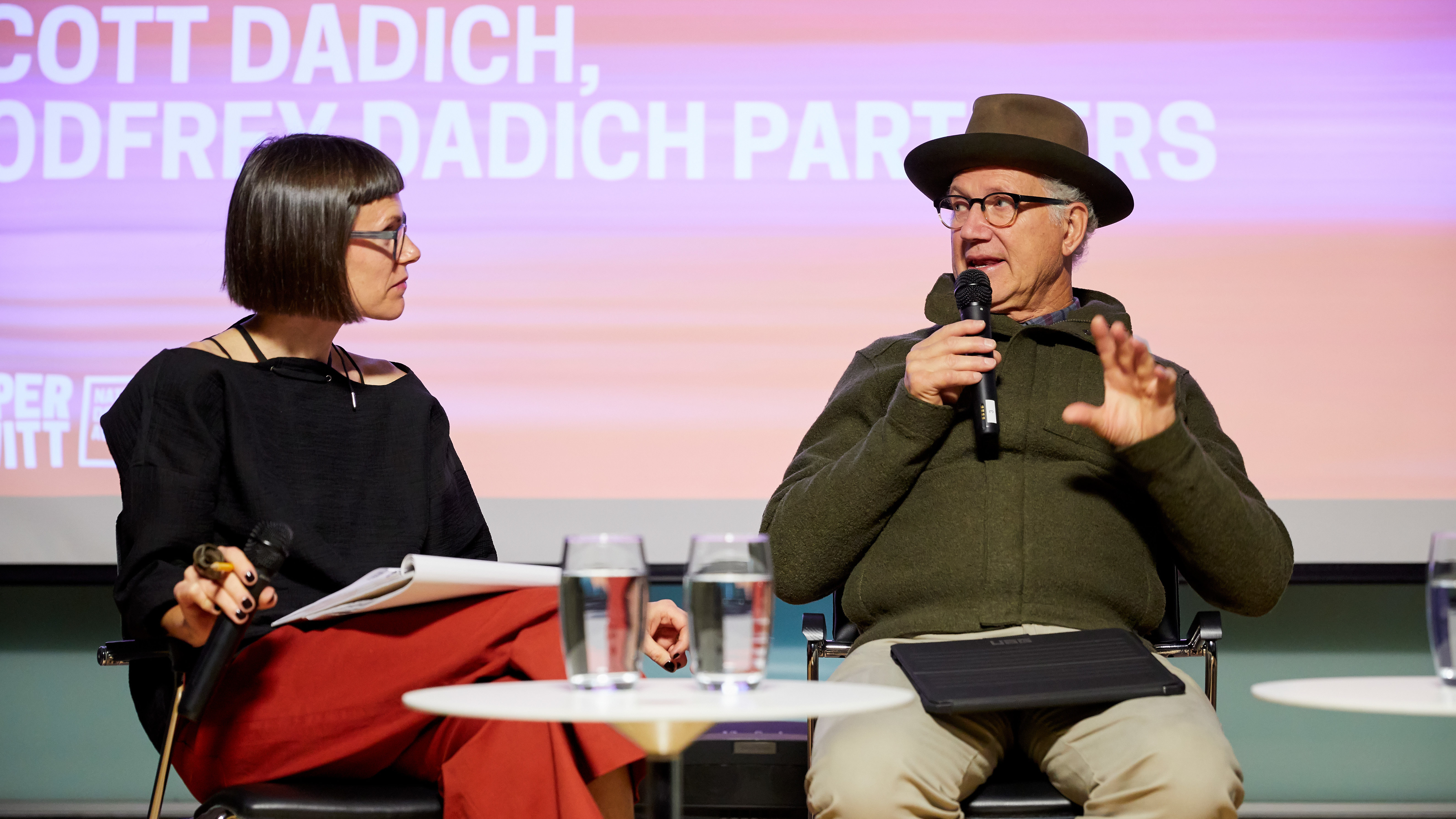 Image of two people on a stage holding microphones. Andrea Lipps has a dark bob haircut and glass, a black shirt and red pants. Tinker Hatfield is on the right, he wears a green woolen jacket and a fedora.