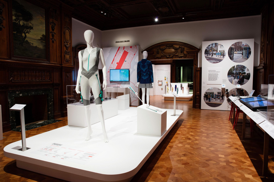 View of the Access+Ability exhibition at Cooper Hewitt. An array of contemporary objects provides a context to the warm, wood-paneled historic gallery. On the center of the plinth are two female mannequins dressed in futuristic garments (The Aura Power Suit, a gray bodysuit with hexagons of a light green material) and the SoundShirt, a navy blue shirt crisscrossed with lighter blue lines). In the distance can be seen ALLELES funky, colorful prosthetic leg covers.