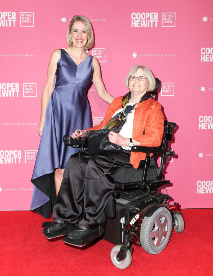 Caroline Baumann, a statueque woman with a blonde bob wearing a flowing purple dress, and Dianne Pilgrim, a wheelchair user with short silver hair wearing an orange cardigan and black pants, pose on the red carpet at the National Design Awards Gala. Behind them is a pink wall printed with the repeating logo of the Cooper Hewitt National Design Awards.