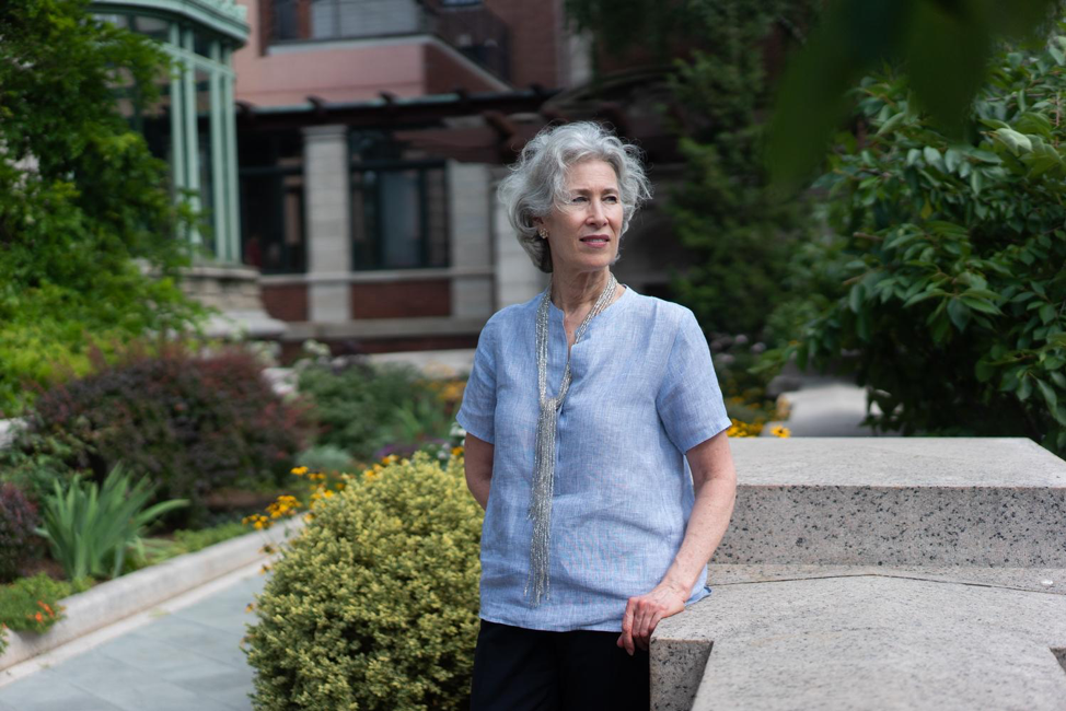 Cara McCarty, an elegant woman with a fair complexion and wind-swept silver hair, gazes thoughtfully into the distance while standing in the Cooper Hewitt Terrace. She is wearing a light blue blouse and a long silver necklace.