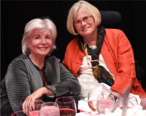 Kay Allaire, a fair-haired woman, pals around with Dianne at the National Design Awards Gala. They are seated at a table with wine glasses on it.