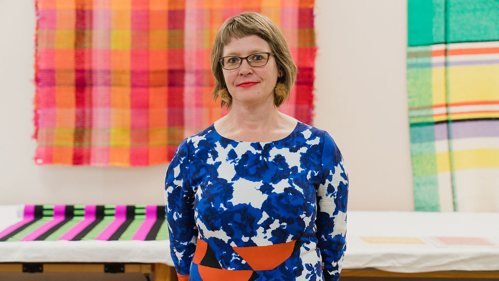 Image of Kate Irvin, who stands in the center of the frame. Brightly colored textiles hang behind her. She wears a bright blue flower print dress, has a short brown bob haircut and wears dark rimmed glasses