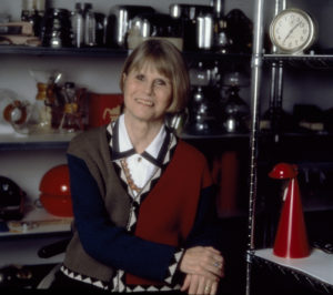 Dianne Pilgrim, seen here in her middle years, is seated in a collections storage area, surrounded by objects such as a clock, a coffeemaker, and a lamp. She has a blonde bob and is smiling. Dianne is a wheelchair user.