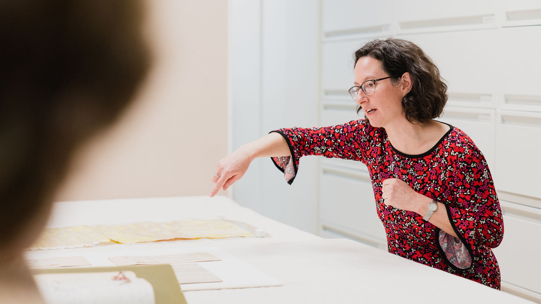 Image of Sarah Vezina, who is pointing at a textile on a table. She has a red flower print dress, dark curly hair to her shoulders, and dark glasses