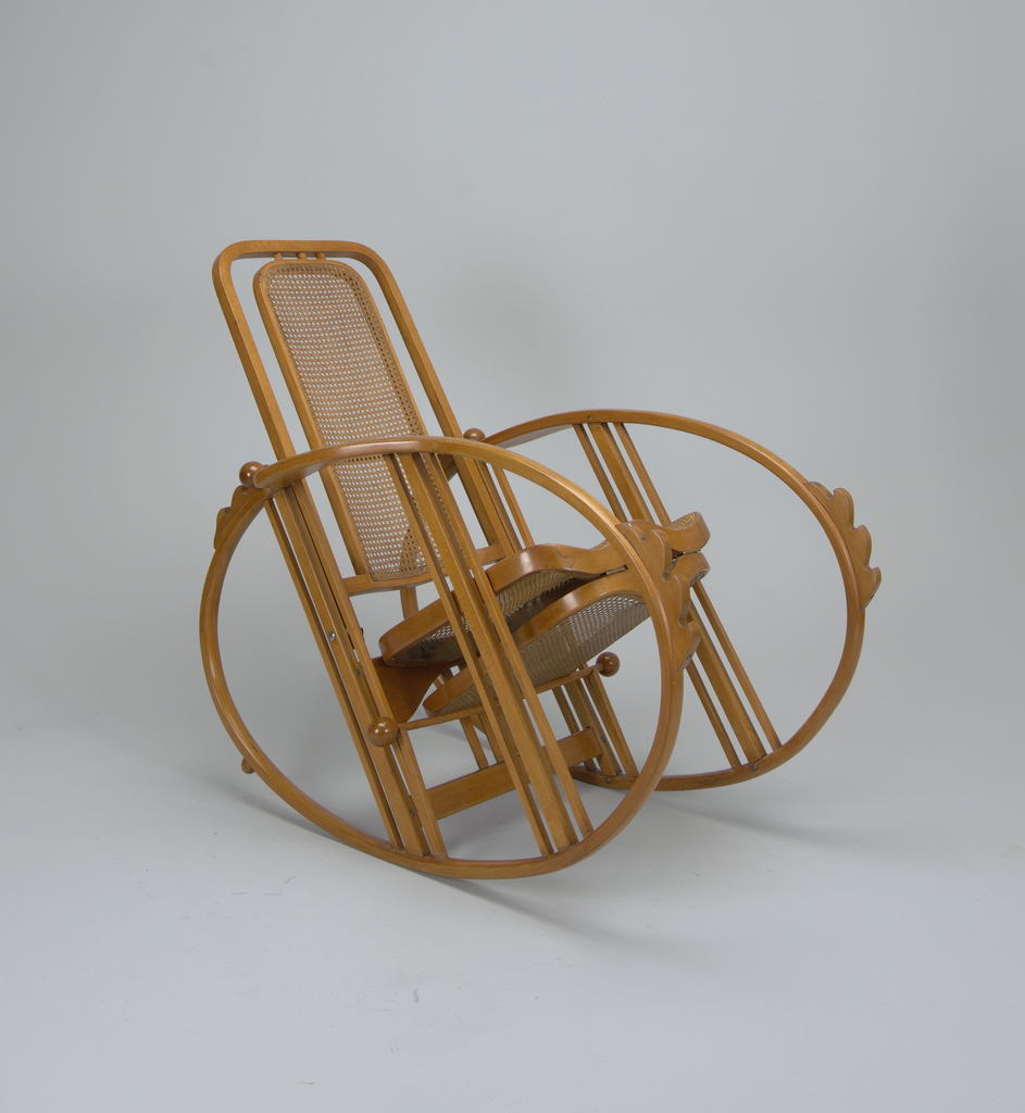 Image features an adjustable reclining rocking chair made of light brown, bent beechwood with woven cane back, seat, and foot rest, hinged to fold under seat. The chair sits on two ovals which serve as its arms and rockers. Please scroll down to read the blog post about this object.