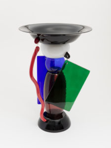 Image features a broad-rimmed circular bowl on a tall base of assembled oval and globular glass forms of opaque or translucent blue, purple, white, and red. The bowl and base decorated with transparent rectangular panels, one blue and one green, and a wavy opaque red glass rod, all hung from brass hooks. Please scroll down to read the blog post about this object.