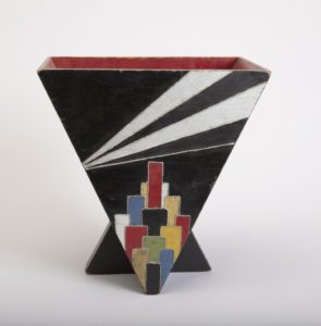 Image features a double-sided wastebasket in the shape of an inverted triangle with two triangular feet and a red interior. One side shows a black background with silver-toned rays emanating from top right corner and a 'cityscape' of colorful overlapping rectangles at the base. Please scroll down to read the blog post about this object.