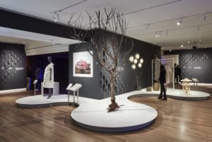 A bare tree stands in the middle of an indoor gallery. On display nearby are objects that appear to be made of natural materials, and a manikin wearing a white dress.